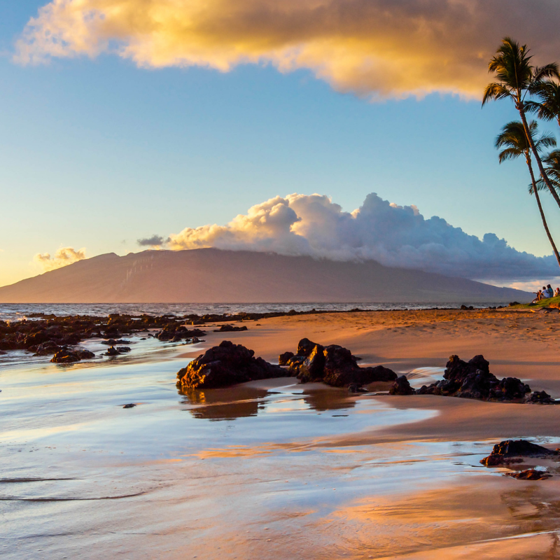 A stunning sunset over a beach in Maui with volcanic rocks and palm trees